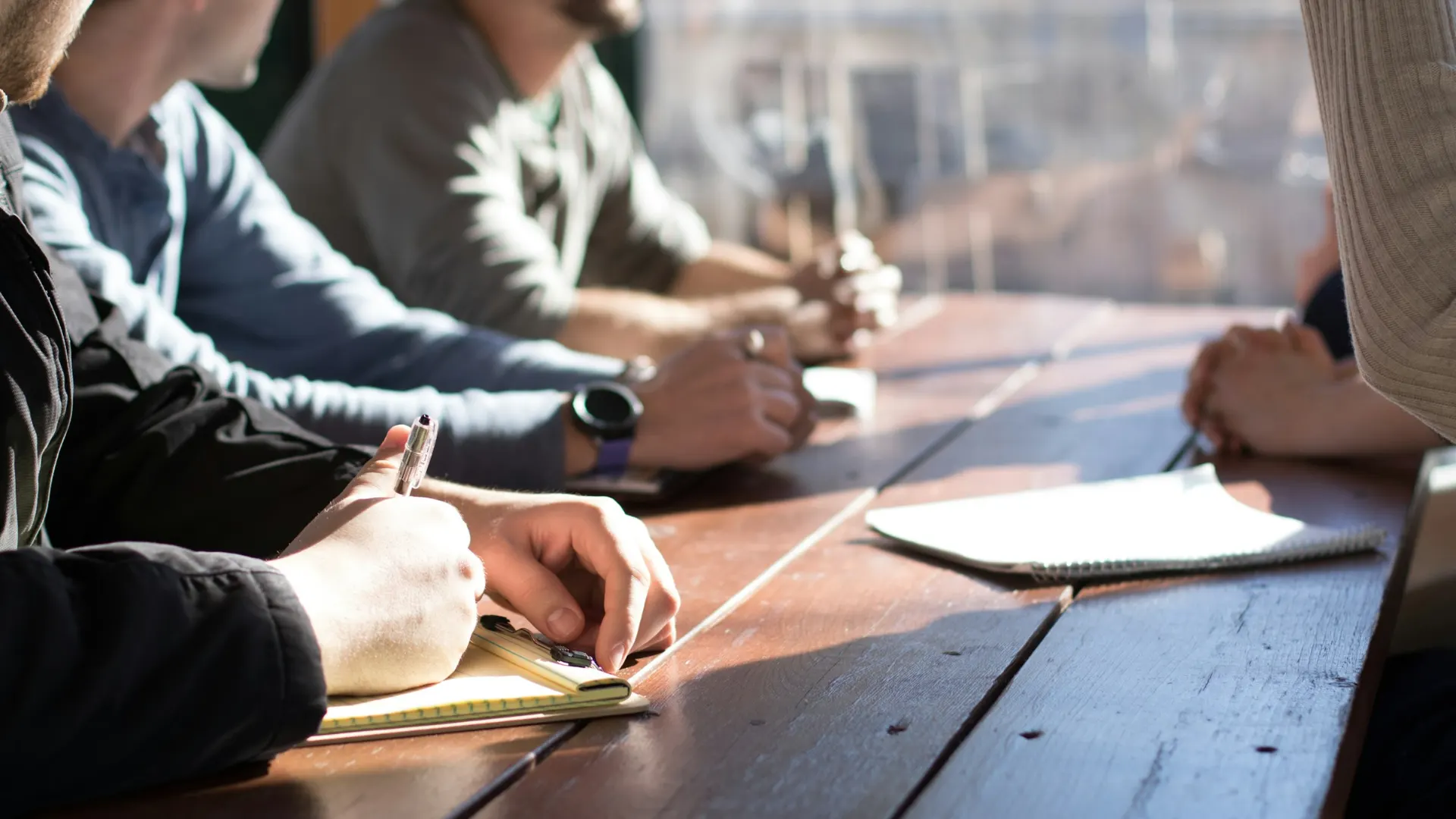 People around a wooden table, one writing notes on a pad.