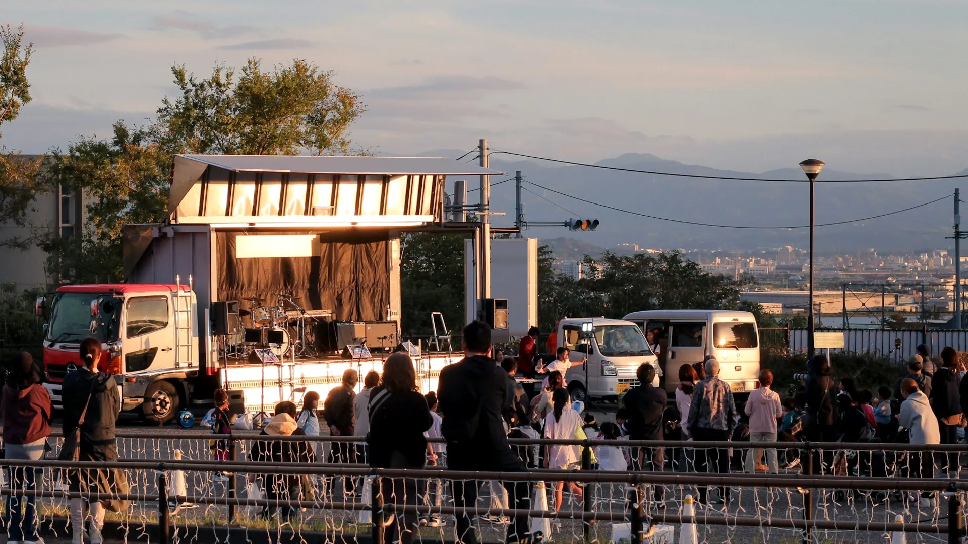 A crowd watching a band on a mobile stage at an outdoor community event, at sunset.